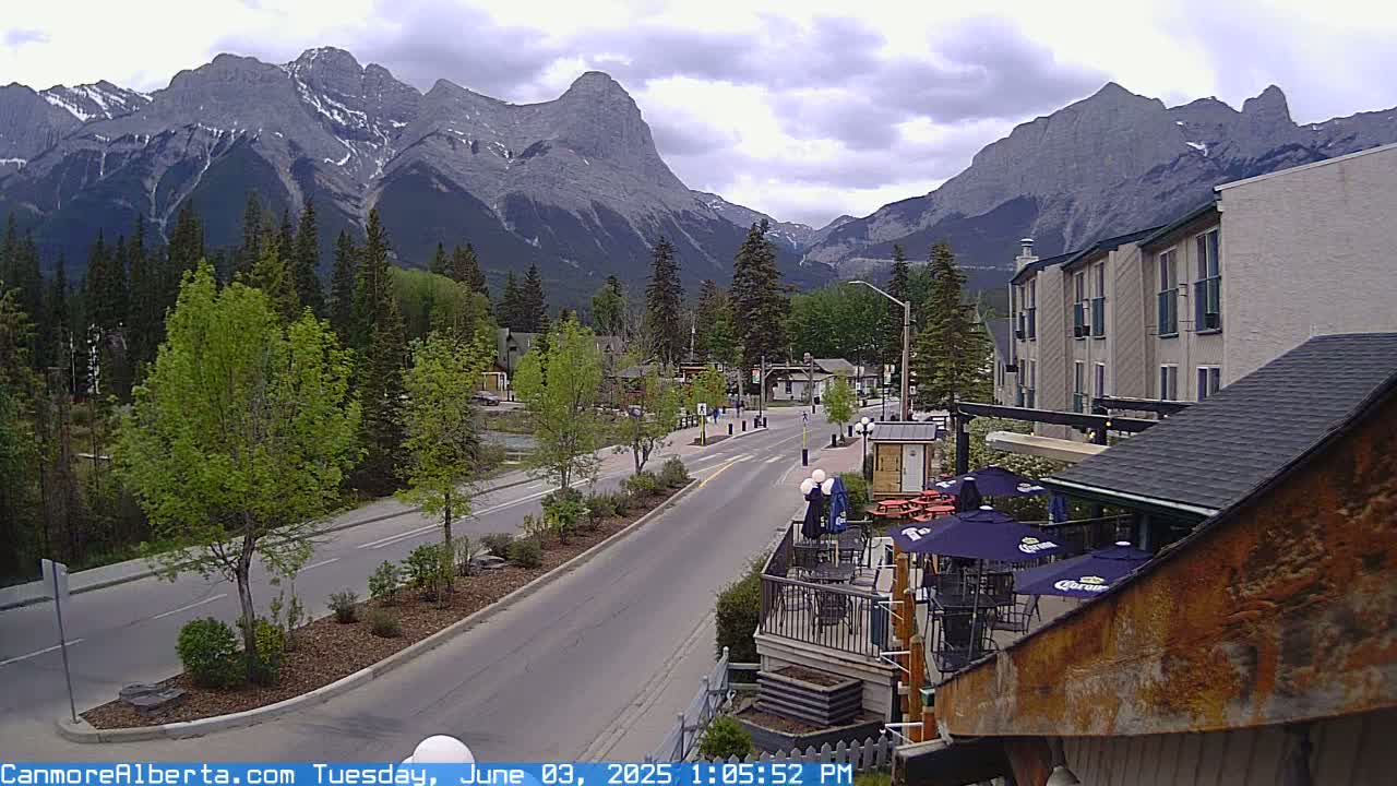 A partly cloudy day reveals a mountain range behind a town's main street, where several buildings, trees, and an outdoor patio are visible.