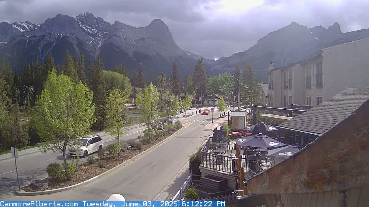 A partly cloudy day reveals a mountain range overlooking a town with a road leading toward it, passing by buildings and a patio area with outdoor seating.