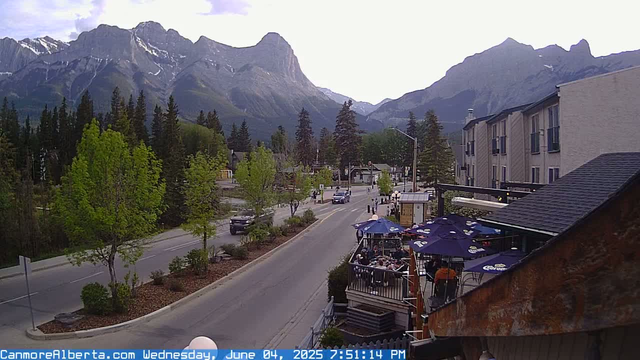 A mountain town street scene shows a partly cloudy evening with cars on a road that leads toward a backdrop of large, imposing mountains, a building with outdoor seating, and green trees.