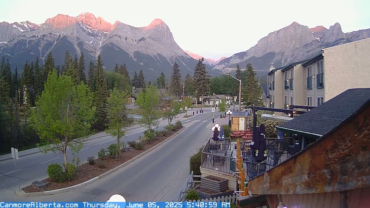 A mountain town street scene at dawn shows a road leading towards majestic, sunlit mountains, with buildings and trees lining the street.