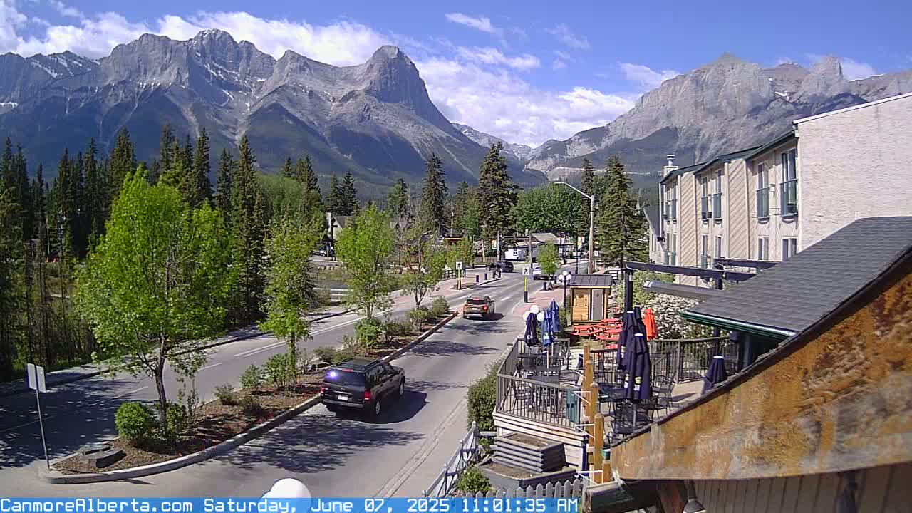A sunny day in a mountain town shows a street scene with cars driving past buildings and outdoor seating, with a large mountain range in the background.