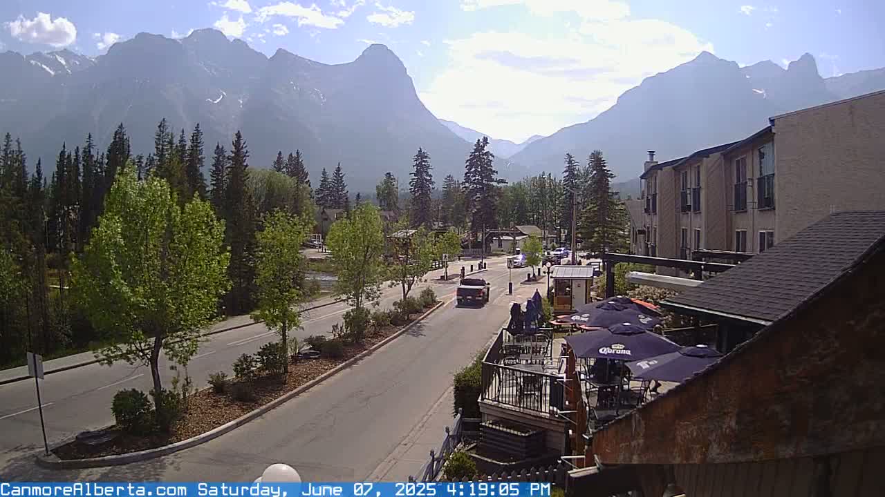 A sunny day reveals a mountain range in the background of a town street scene with cars driving on a road lined with trees, and a patio with umbrellas in the foreground.