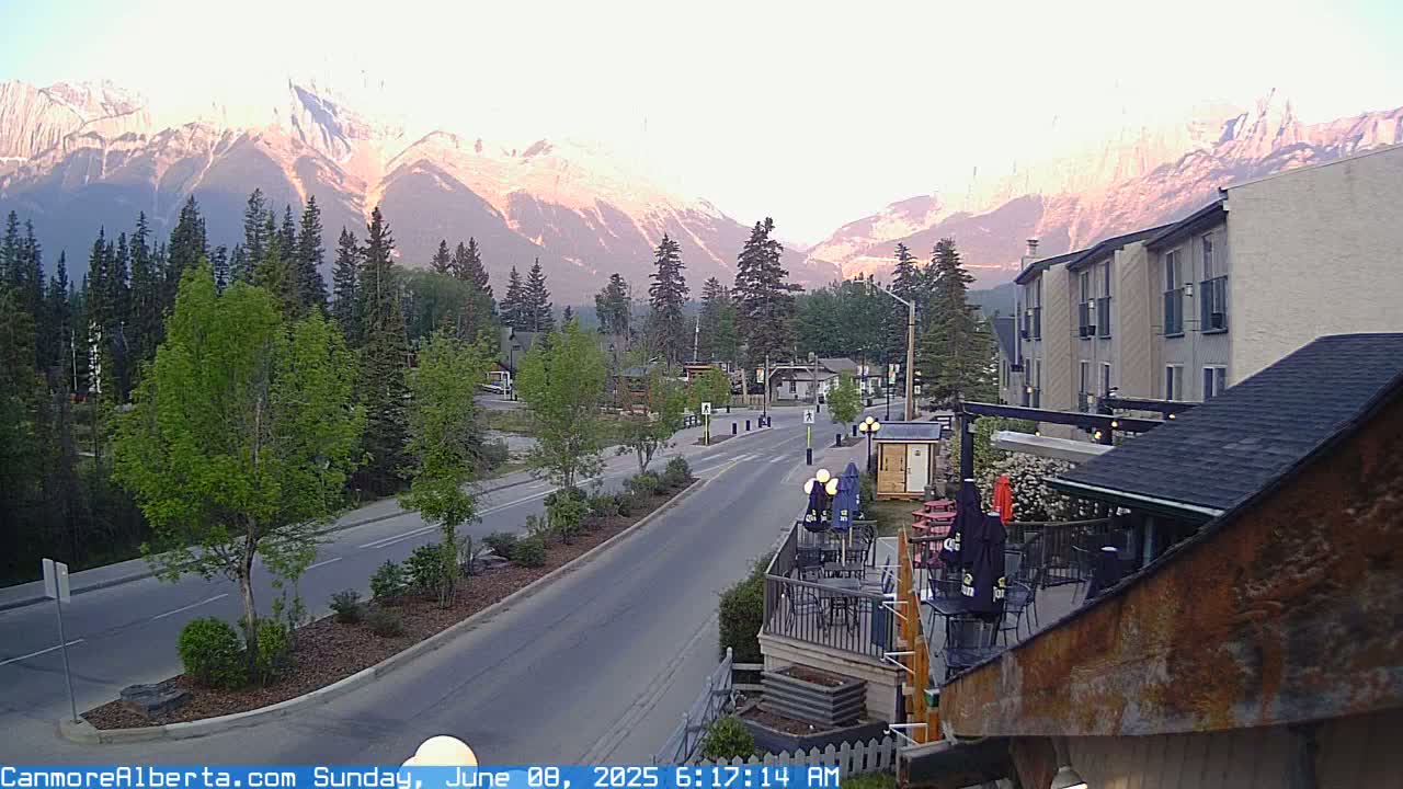 A mountain range glows pink and orange at sunrise over a quiet town street lined with trees and buildings, with a patio visible in the foreground.