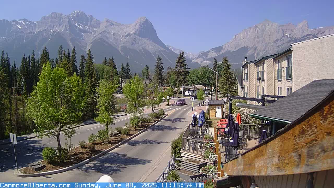 A sunny mountain town scene shows a road leading toward a backdrop of majestic mountains, with trees lining the street and a patio area visible in the foreground.