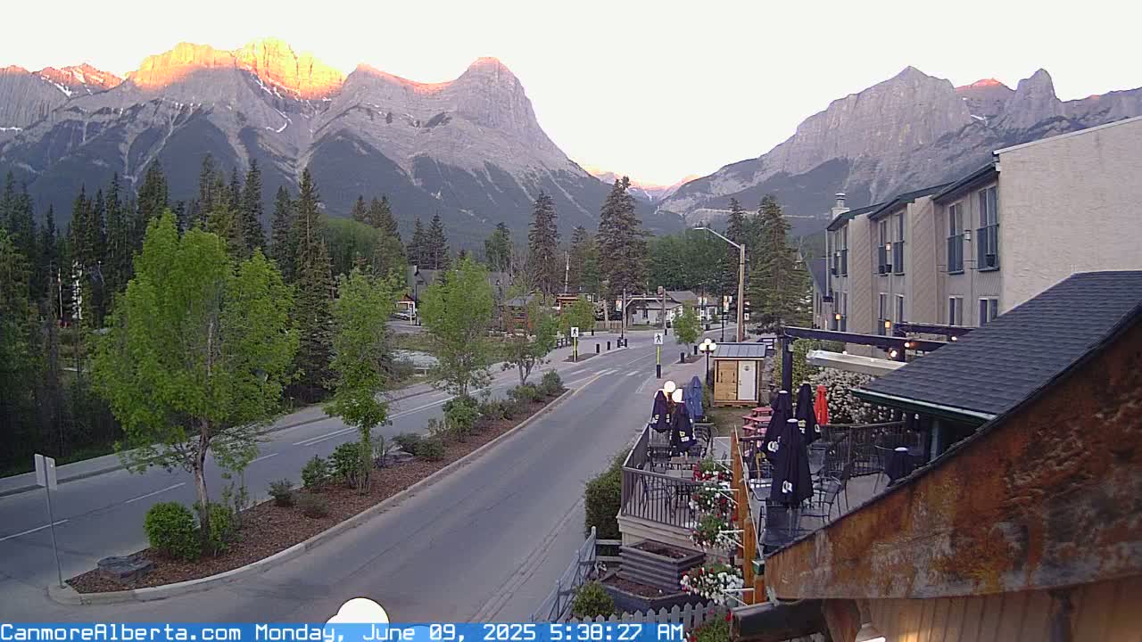 A mountain town street scene at sunrise, showing a road leading towards majestic, sunlit mountains in the background.