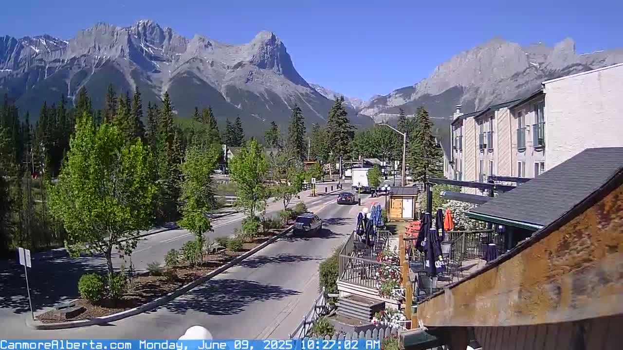A sunny mountain town scene shows a paved road with cars driving past a restaurant patio, all set against a backdrop of large, rocky mountains.
