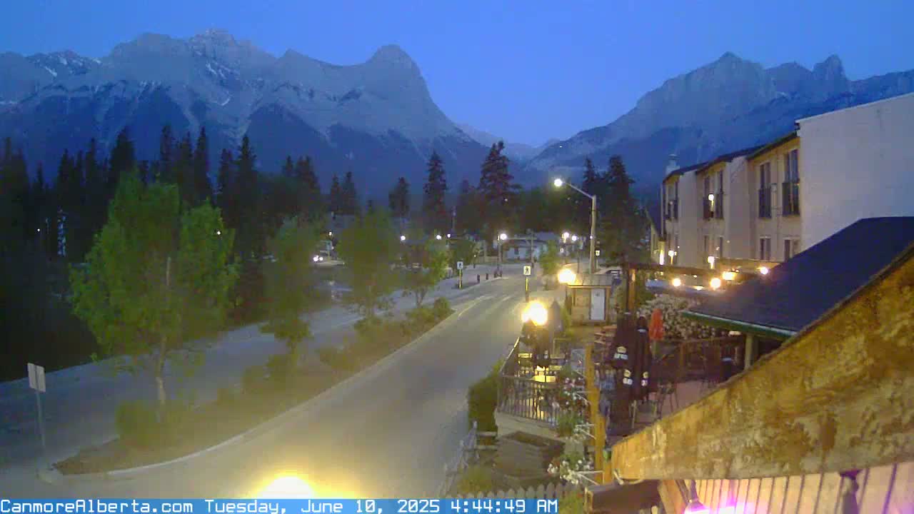 A dimly lit street curves past buildings towards snow-capped mountains under a twilight sky.