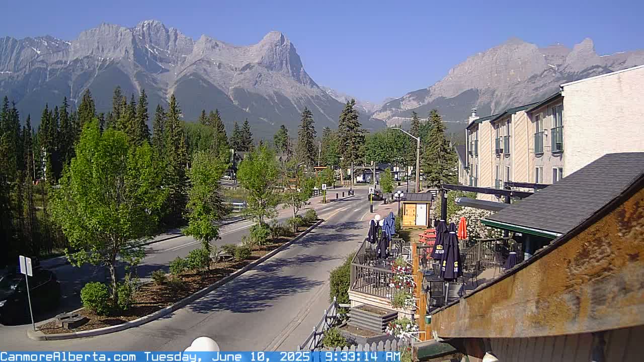 A town street scene featuring a multi-story building, outdoor patio seating, and a mountain range under a clear blue sky.