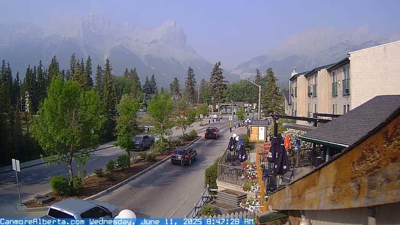 A hazy, sunny day reveals a mountain range in the background, overlooking a street scene with cars and buildings, including a multi-story hotel and a patio area with umbrellas.
