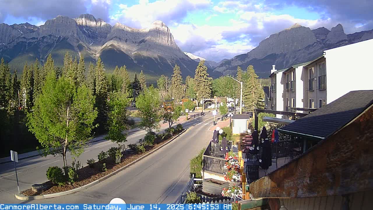 A partly cloudy morning reveals a mountain range behind a town's main street with buildings, trees, and a patio area.