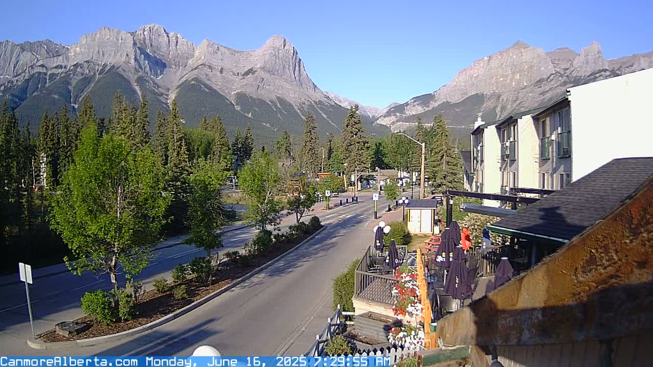 A sunny day reveals a mountain range behind a town with a quiet street, trees, and a patio with umbrellas.