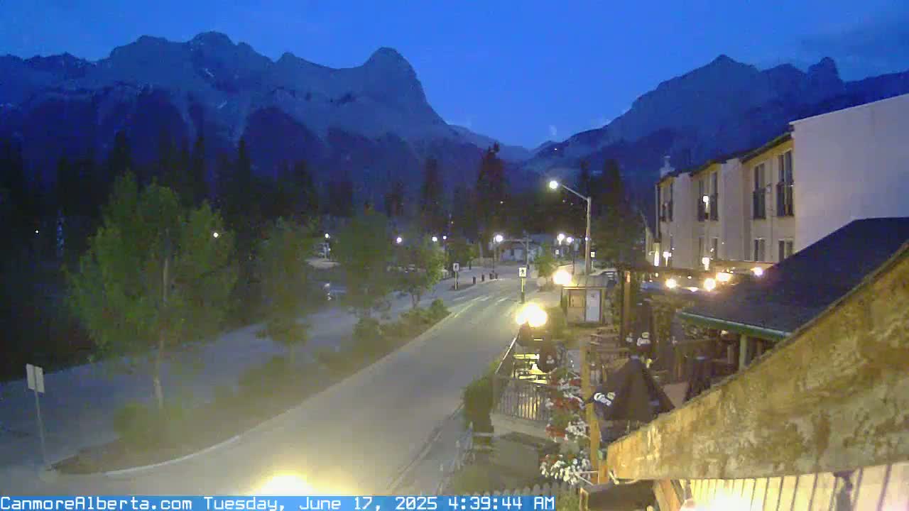 A nighttime view of a town nestled against a mountain range under a twilight sky, with a road curving through the foreground.