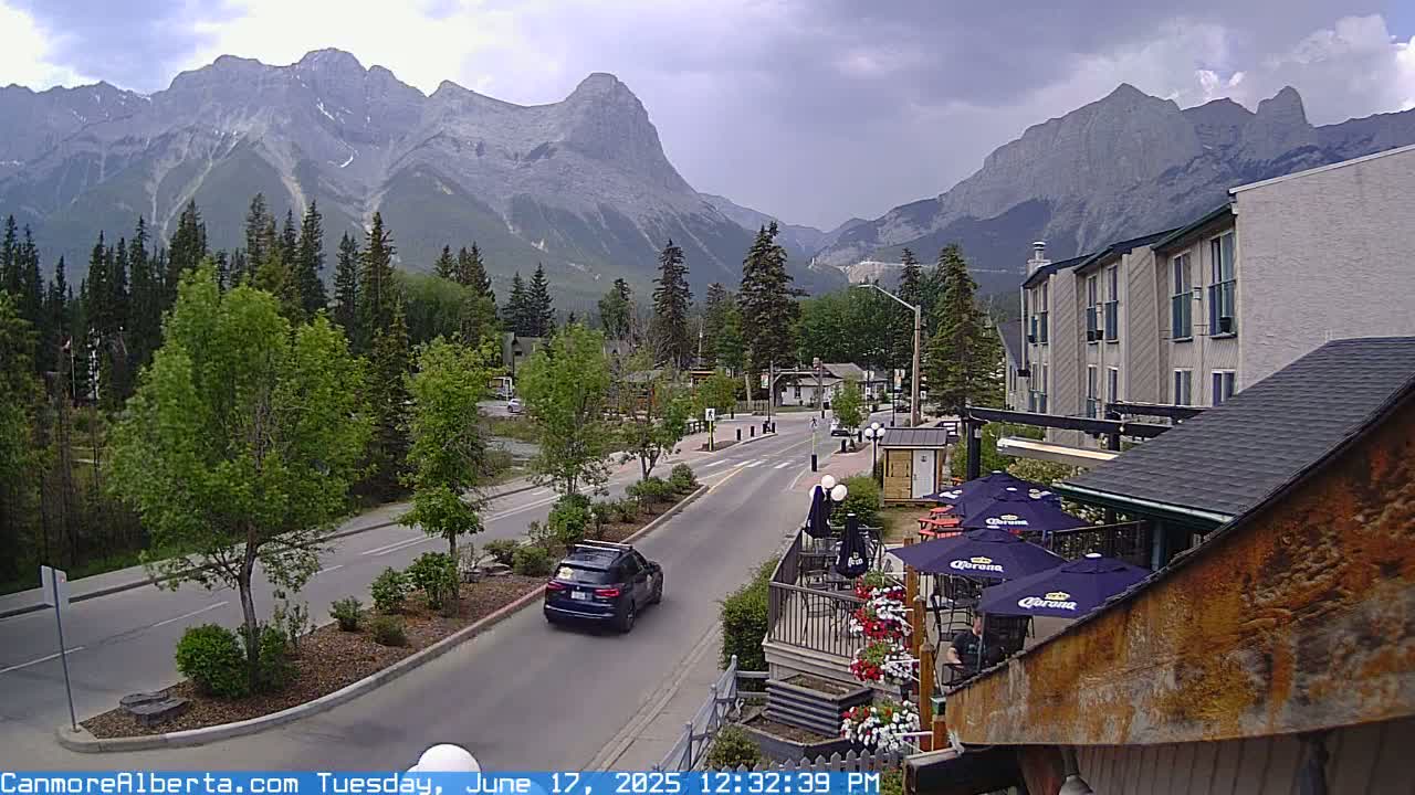 A partly cloudy day reveals a mountain range behind a town's street scene, which includes a multi-story building and an outdoor patio area with umbrellas.