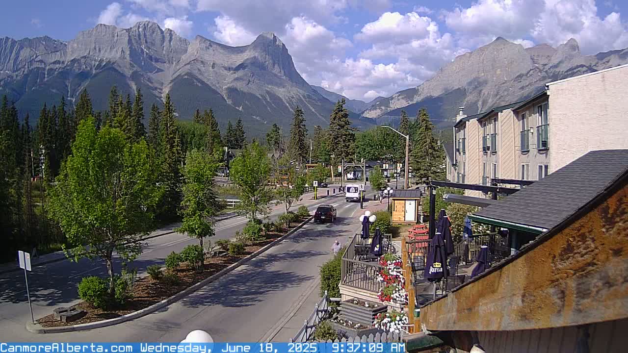 A partly sunny day reveals a mountain range behind a town's street scene, with cars driving along a road lined with trees and buildings.