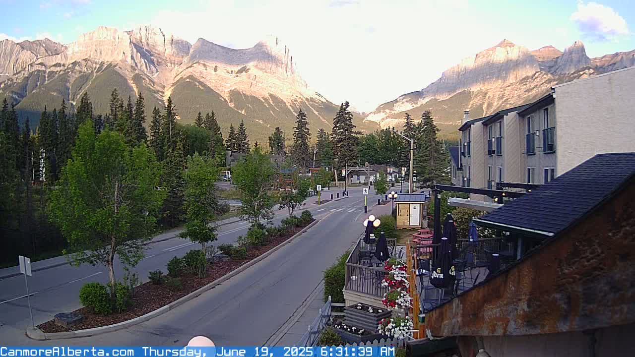 A mountain town street scene at sunrise, showing a road leading towards towering, sunlit mountains, with buildings and trees lining the street.