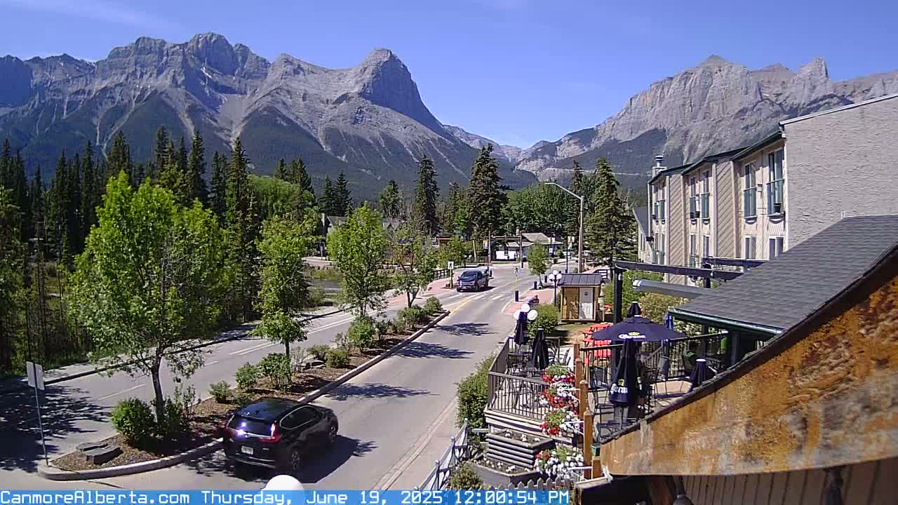 A sunny mountain town scene shows a road with cars, a multi-story building, outdoor seating at a restaurant, and a backdrop of majestic mountains covered in trees.