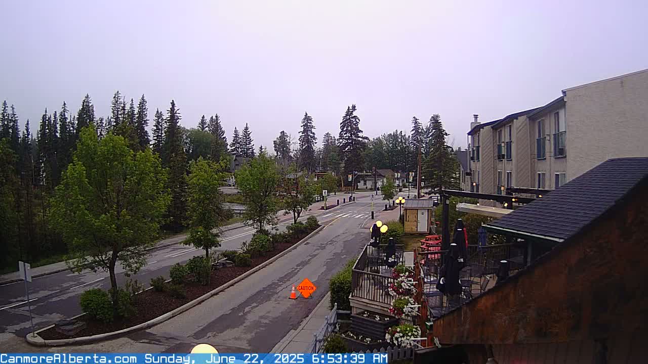 A mostly cloudy day reveals a quiet street scene in a town surrounded by evergreen trees, with a multi-story building and outdoor patio visible in the foreground.