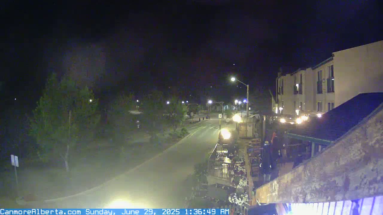 A nighttime, slightly hazy view of a quiet street with a few cars and a multi-story building partially visible, trees lining the road.
