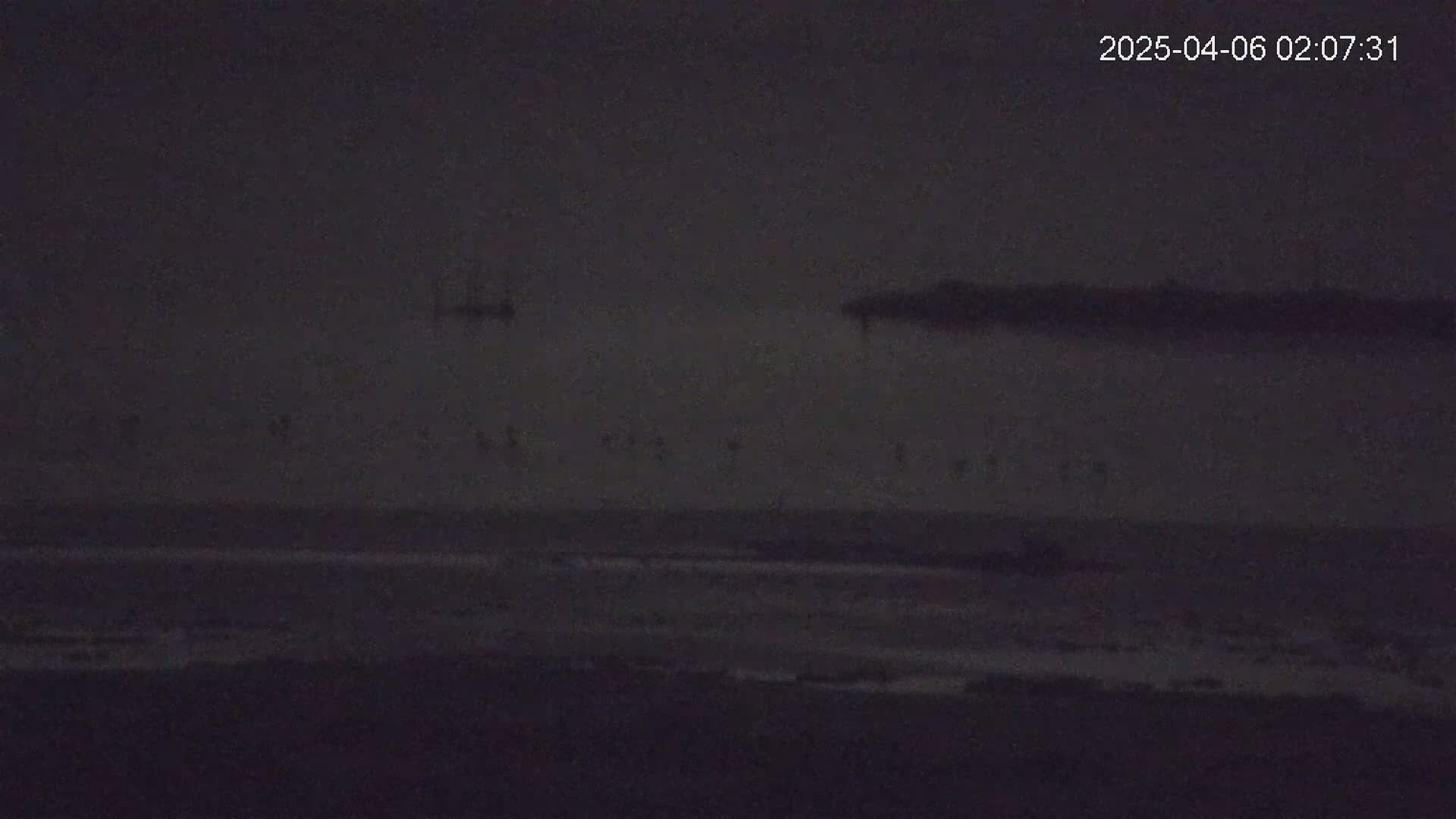 A dark, nighttime scene shows a calm body of water with a distant shoreline under a cloudy sky.