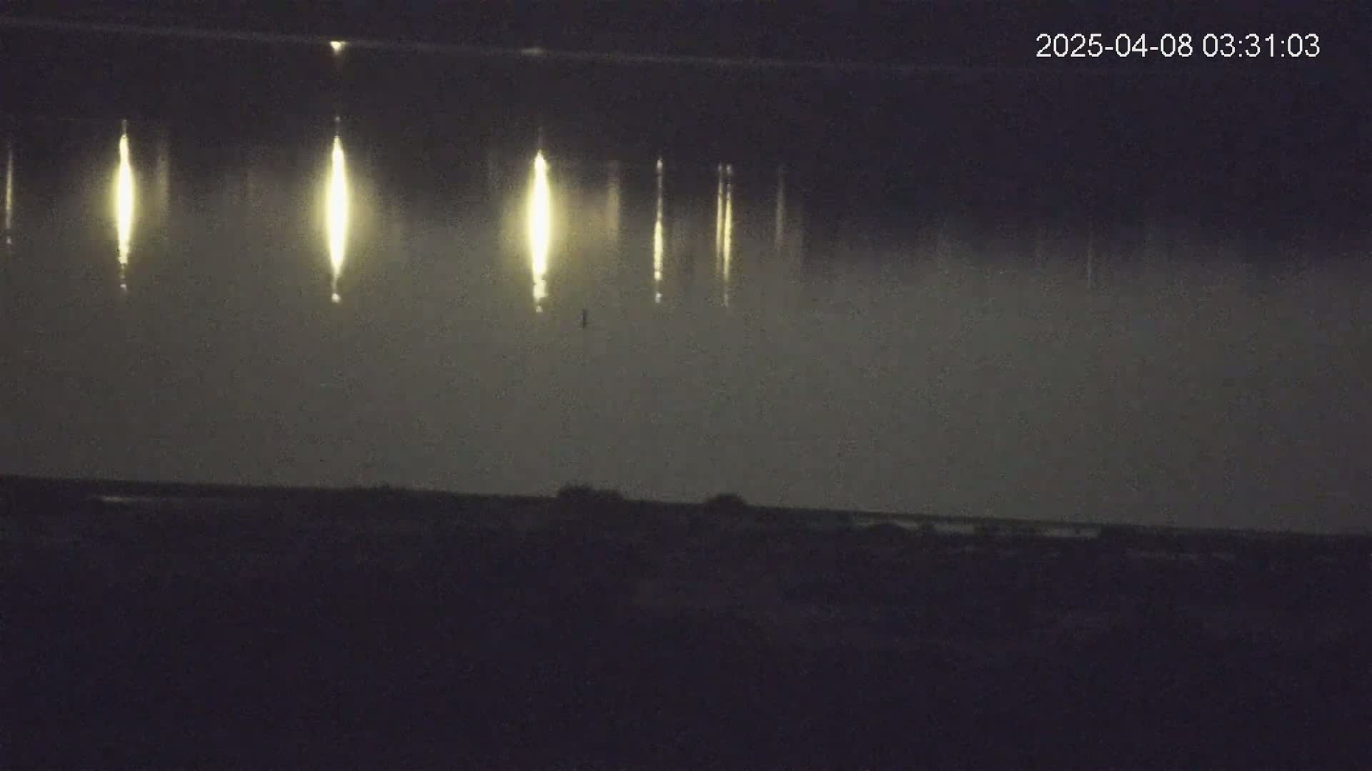 Several vertical streaks of bright light appear over a dark, foggy body of water at night, with a dark shoreline in the foreground.