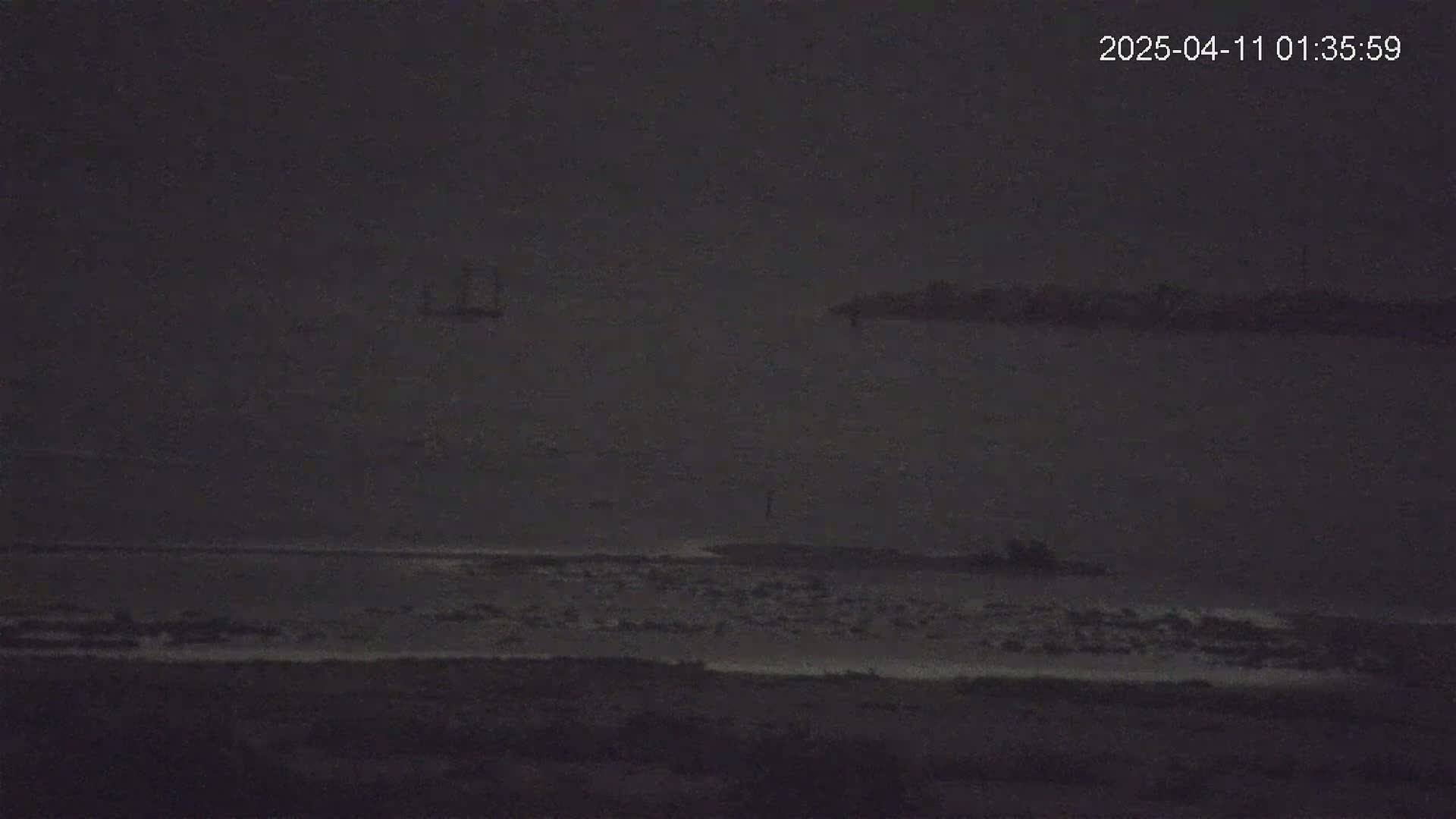 A dark, nighttime view of a calm body of water with a distant shoreline and a small vessel.