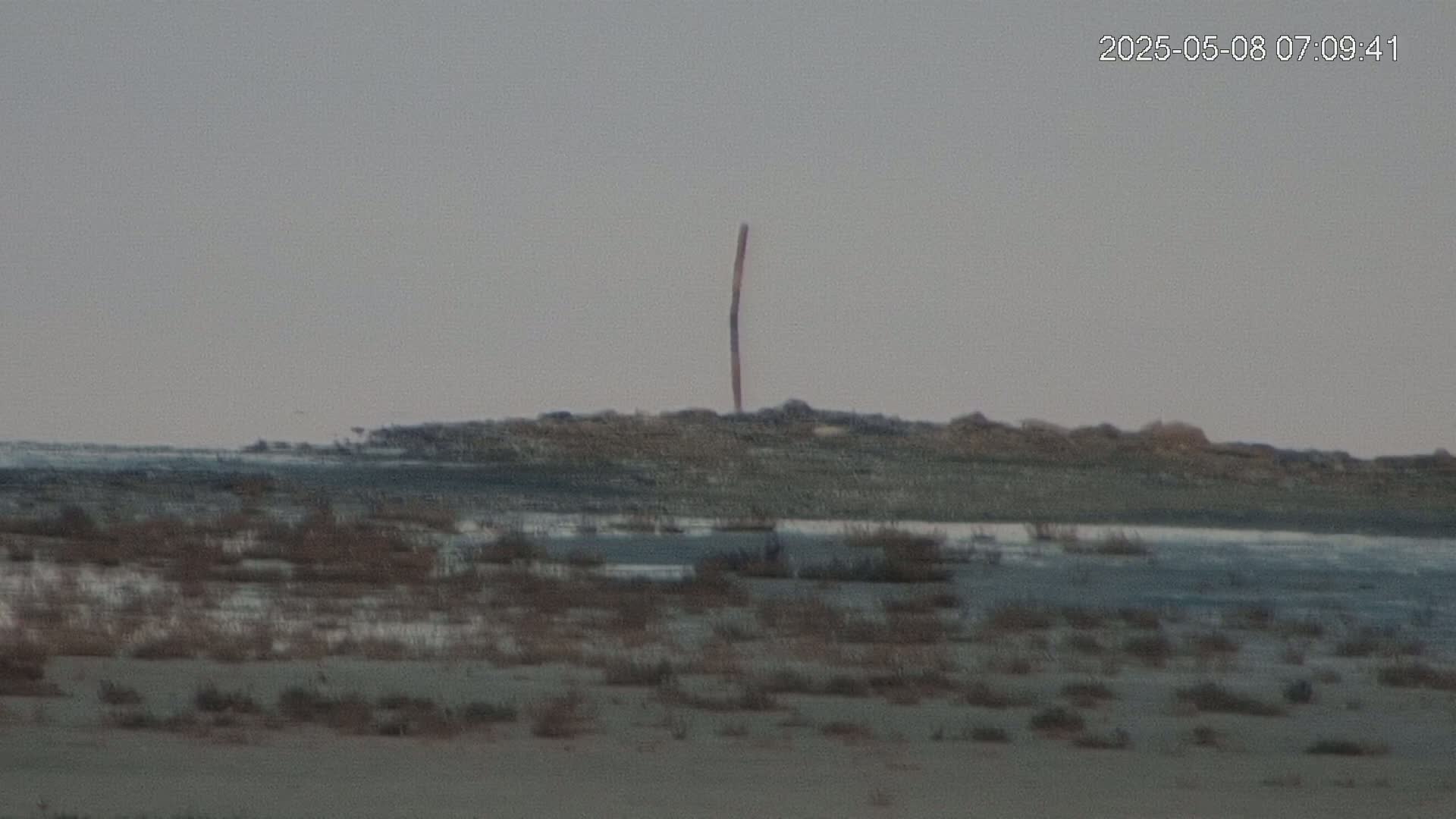 A tall, thin, dark pole stands on a flat, rocky area next to a body of still, grayish water, under an overcast sky.