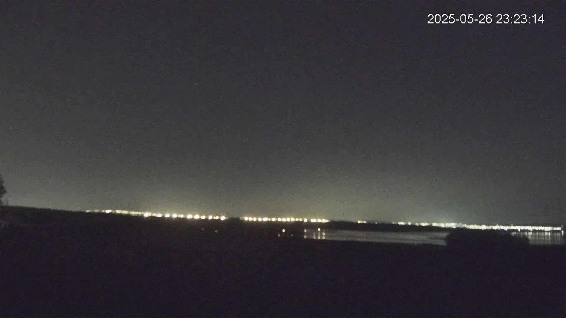 A nighttime view shows a distant line of city lights across a dark body of water under a clear, dark sky.