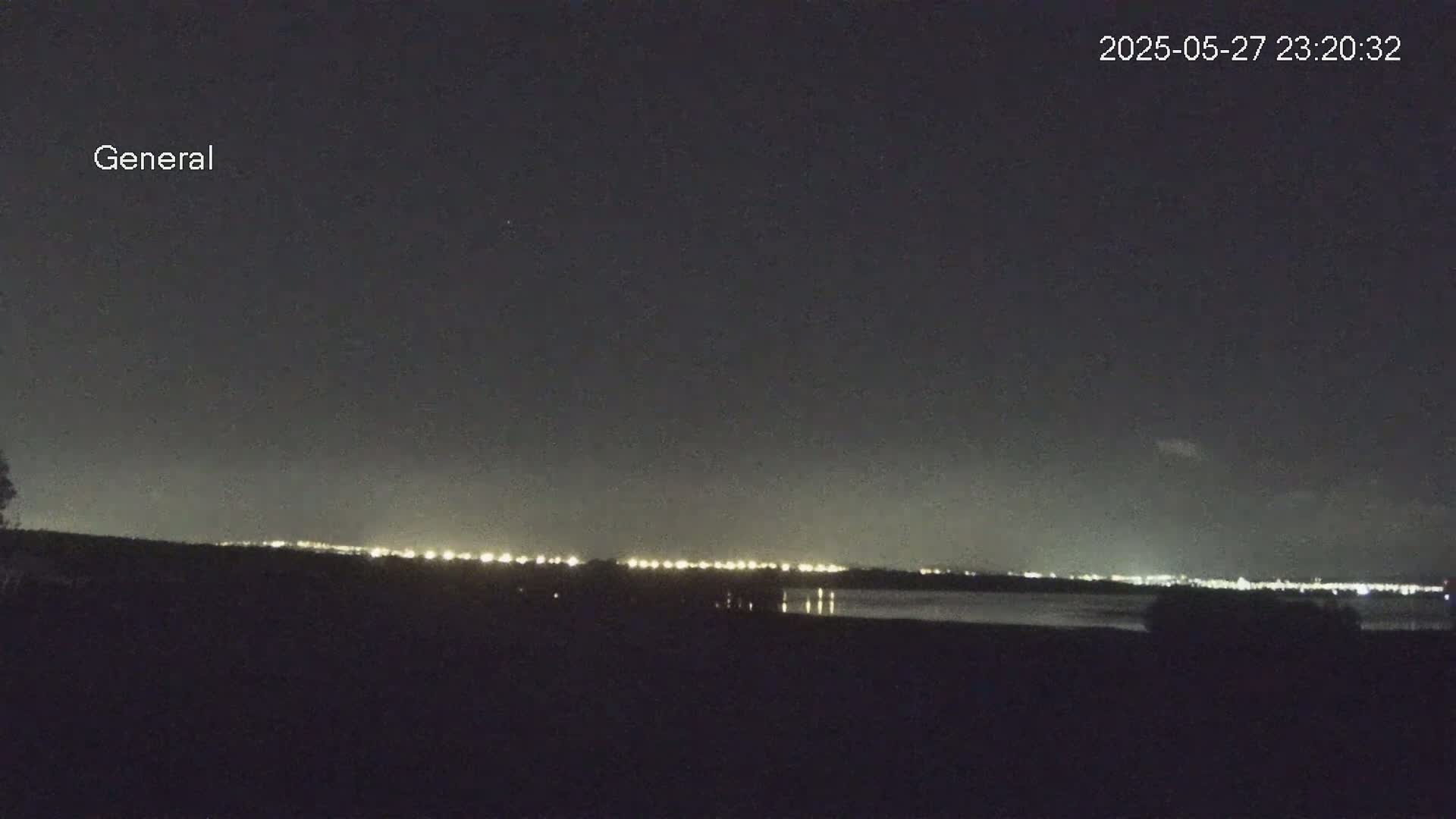 A nighttime view shows a distant line of city lights across a body of water under a dark, clear sky.