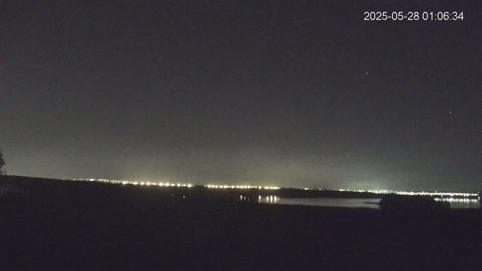 A nighttime view shows a dark landscape with a distant line of lights reflecting on a body of water under a clear, star-studded sky.