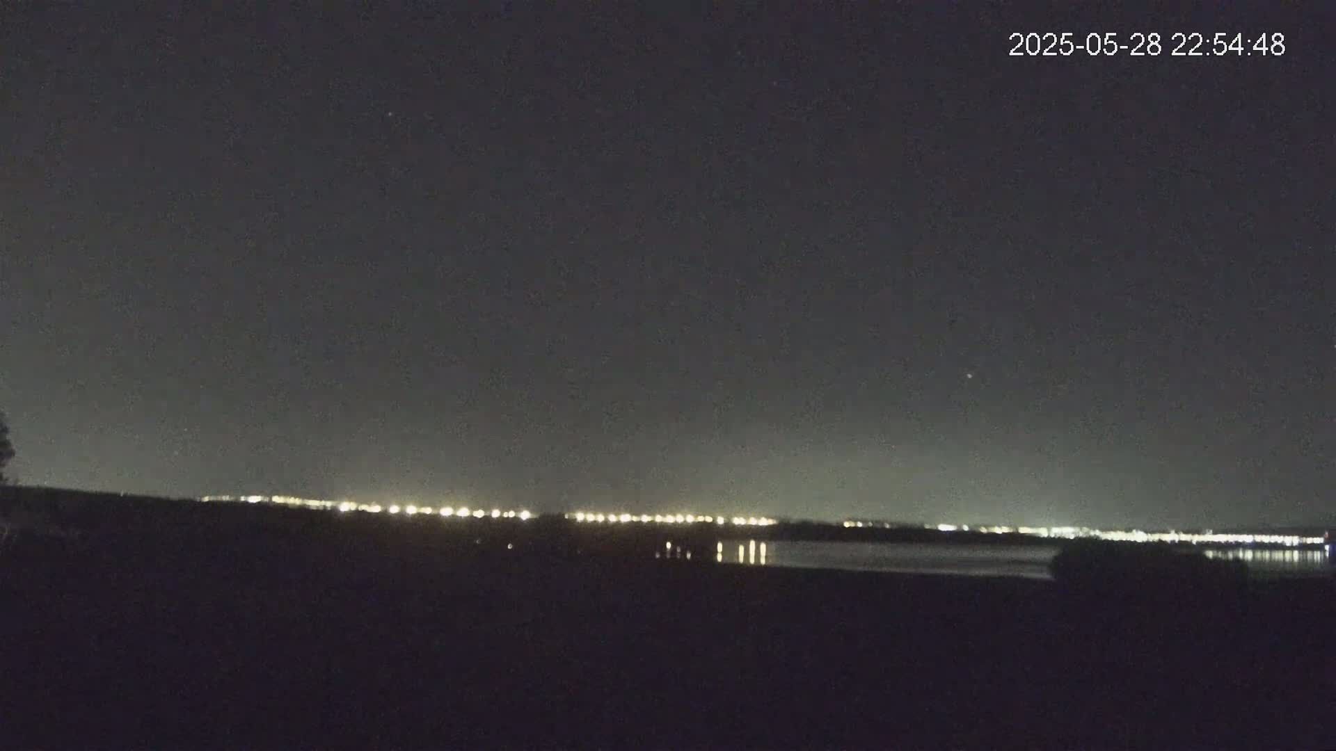 A nighttime view shows a dark body of water reflecting a distant line of city lights under a clear, dark sky.