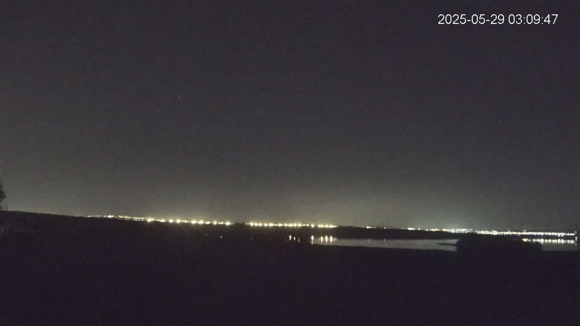 A nighttime view of a distant city skyline illuminated across a dark body of water under a clear, star-filled sky.