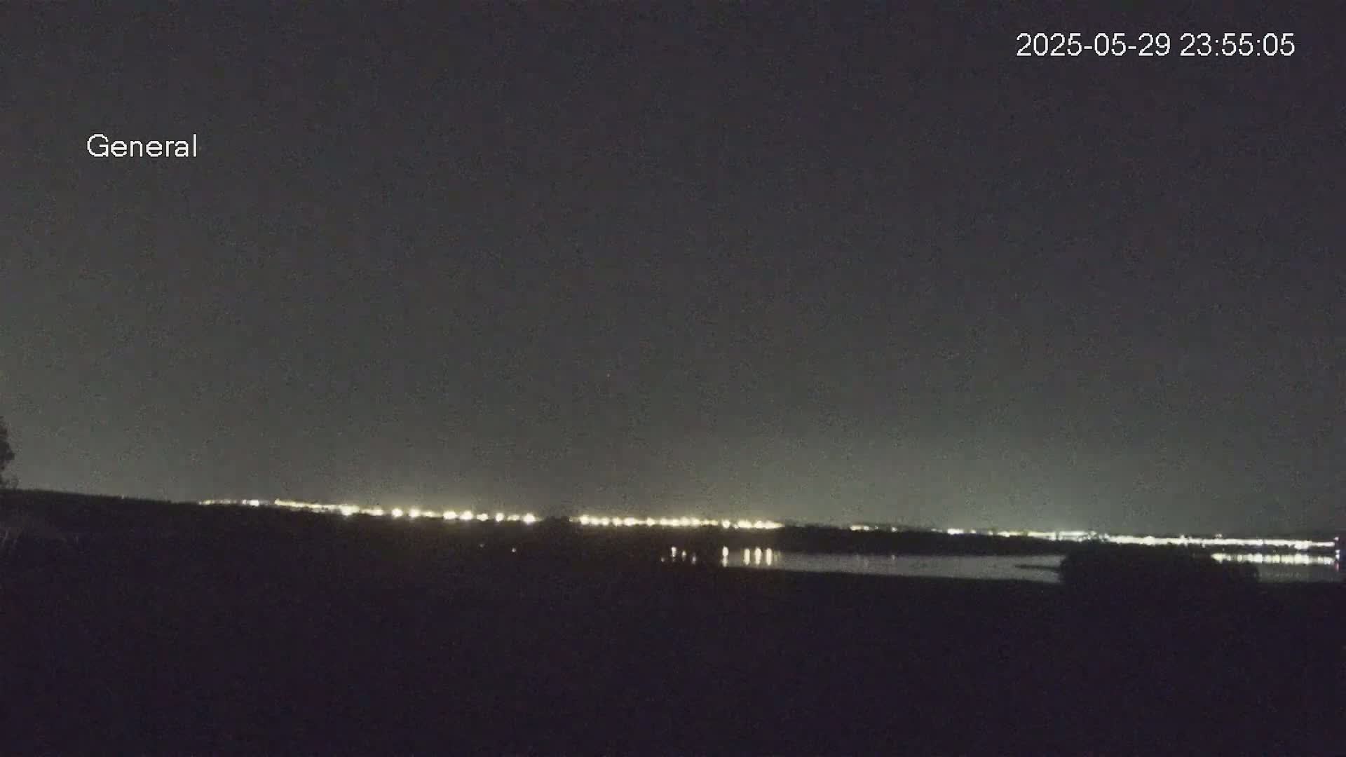 A nighttime view shows a dark, flat landscape with a distant line of lights reflecting on a body of water under a clear, dark sky.