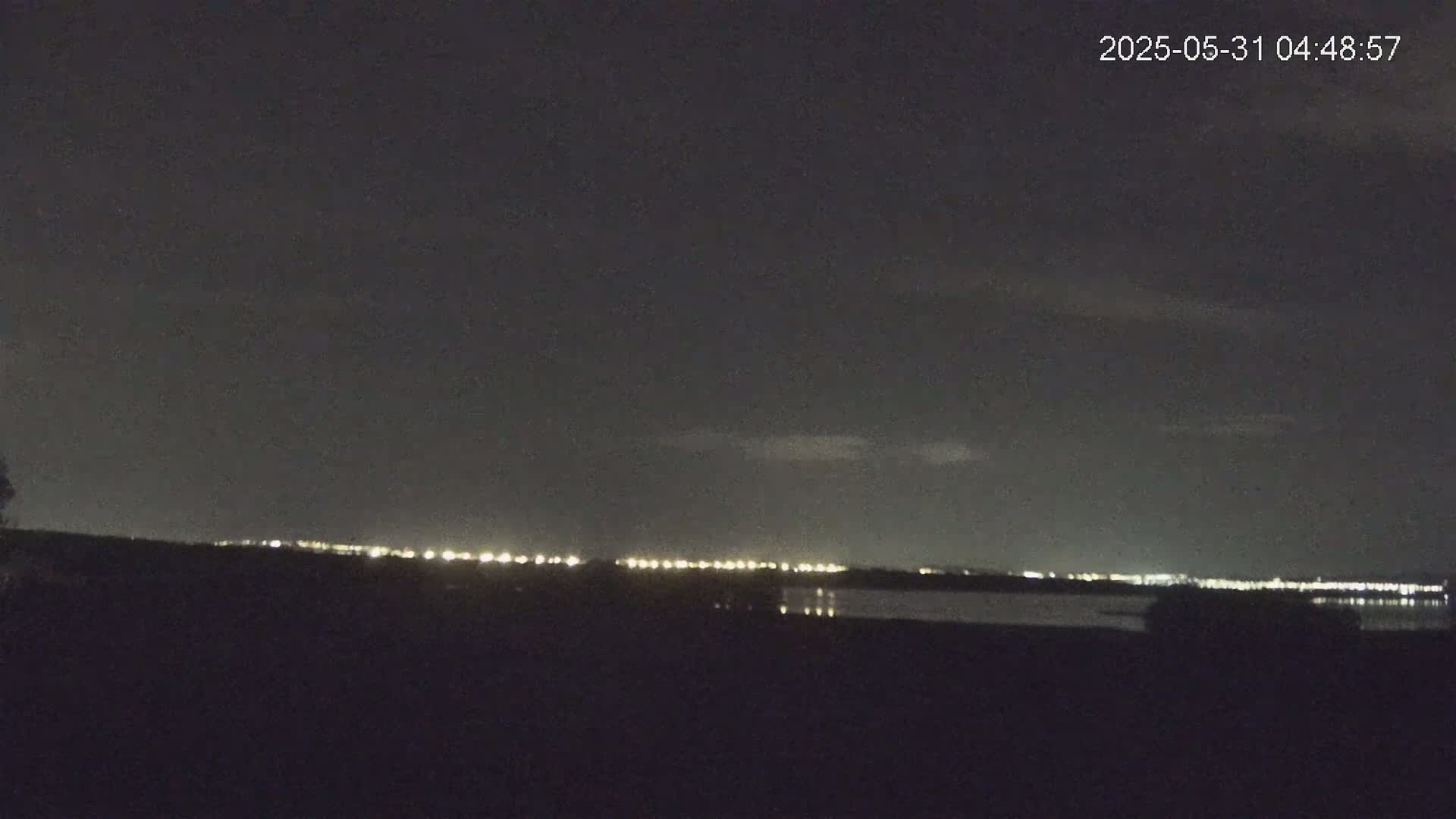 A nighttime view shows a dark landscape with a distant line of lights reflecting on a body of water under a mostly clear, dark sky.