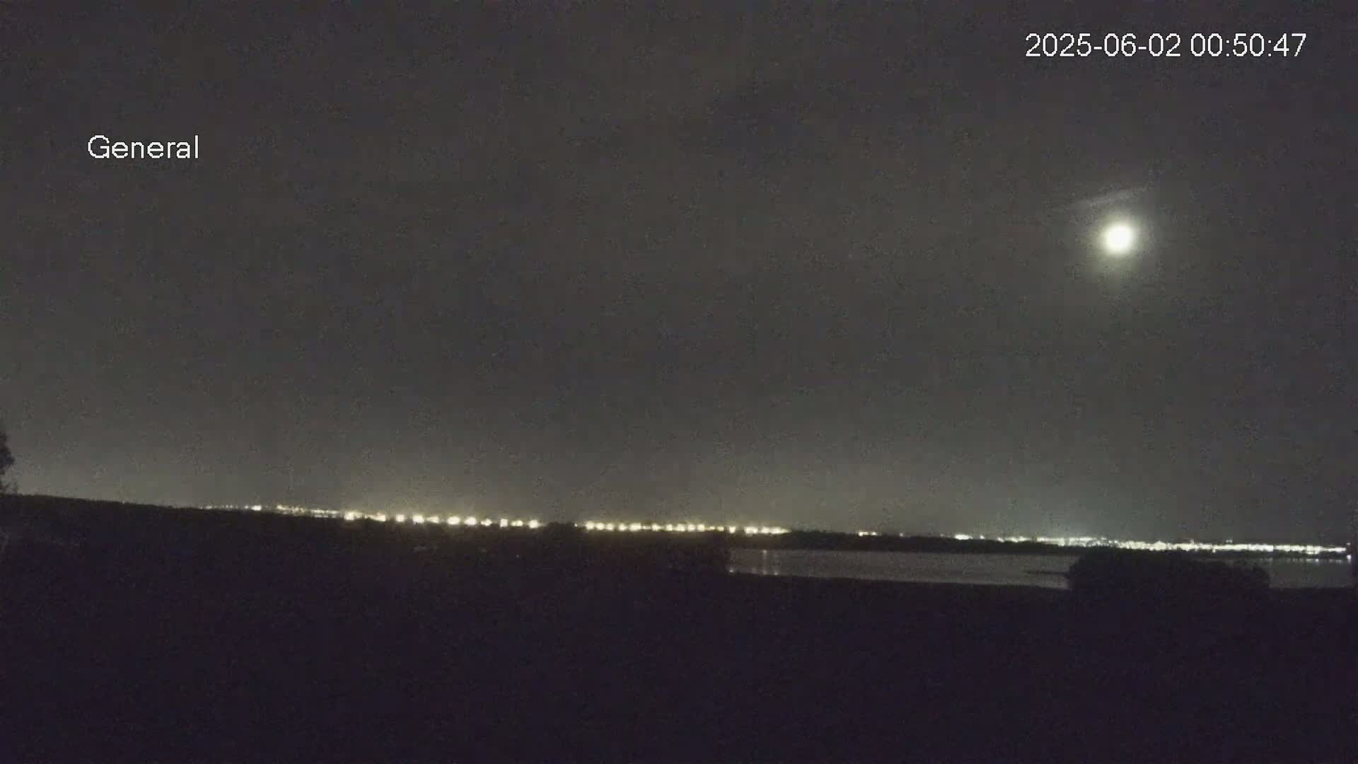 A nighttime view shows a bright moon in the sky above a dark landscape with a distant line of city lights reflecting on a body of water.