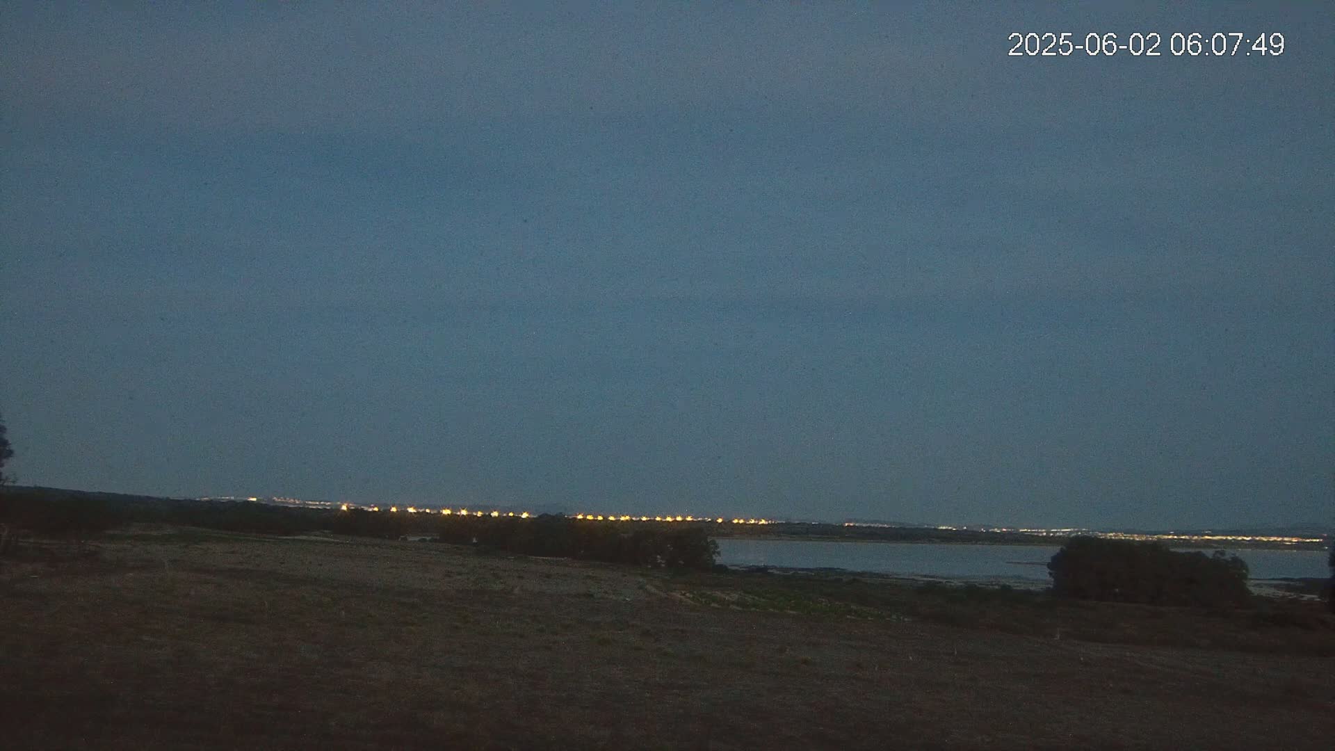 A dark, flat landscape at twilight shows a line of distant lights across a body of water.