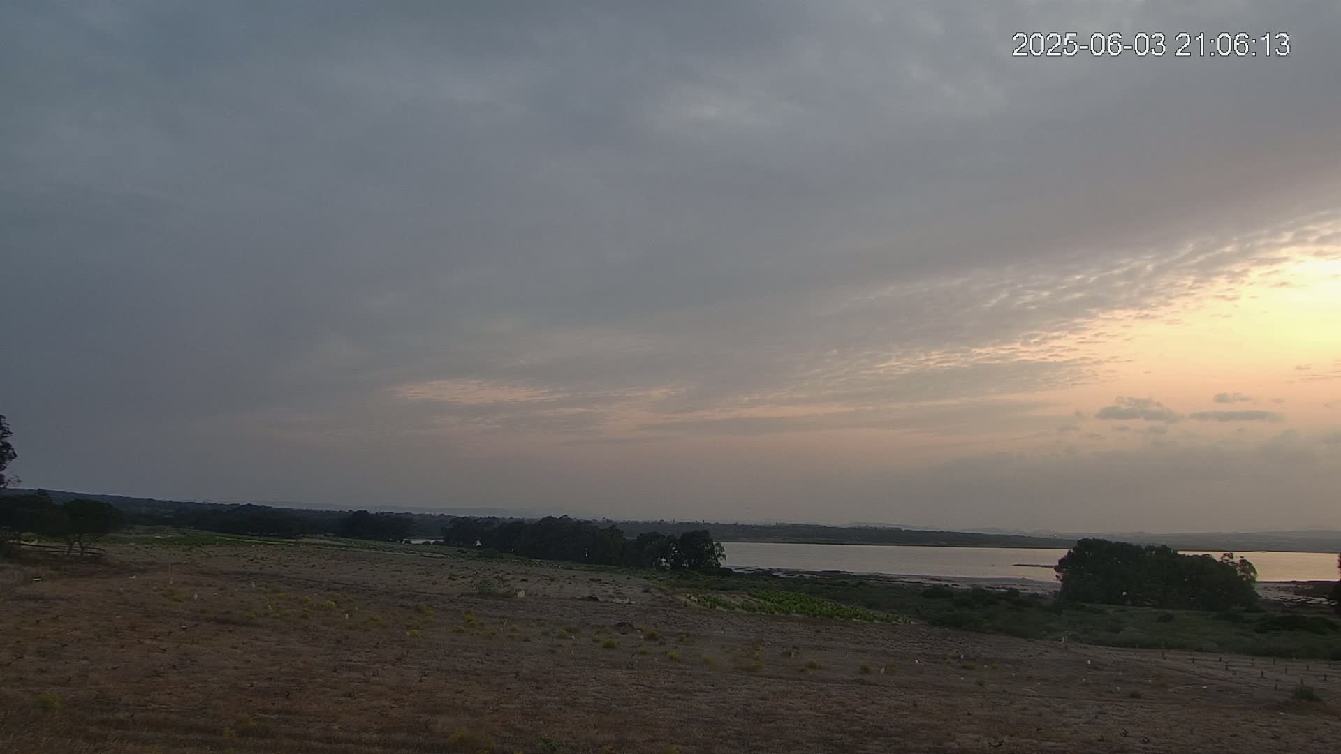 A mostly cloudy sunset over a flat, dry landscape with a calm body of water in the distance.