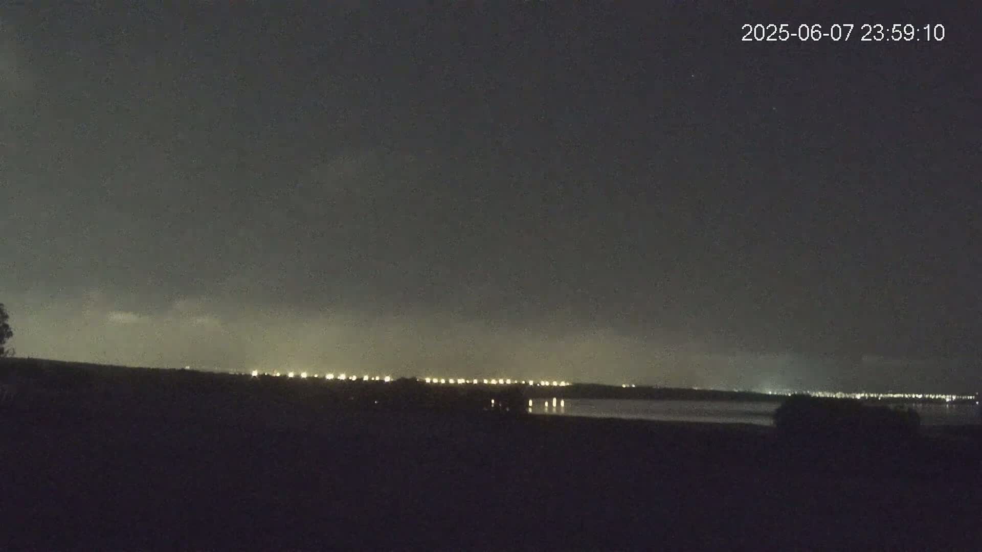 A nighttime view shows a distant line of lights across a dark body of water under a mostly clear, dark sky.
