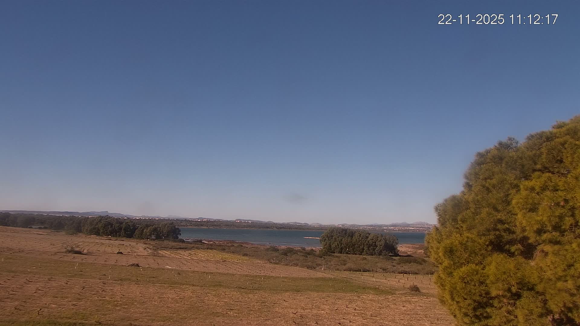 A clear blue sky hangs over a sunny landscape featuring dry fields and green trees in the foreground, with a large body of water bordered by distant land and faint mountains under a bright sun.