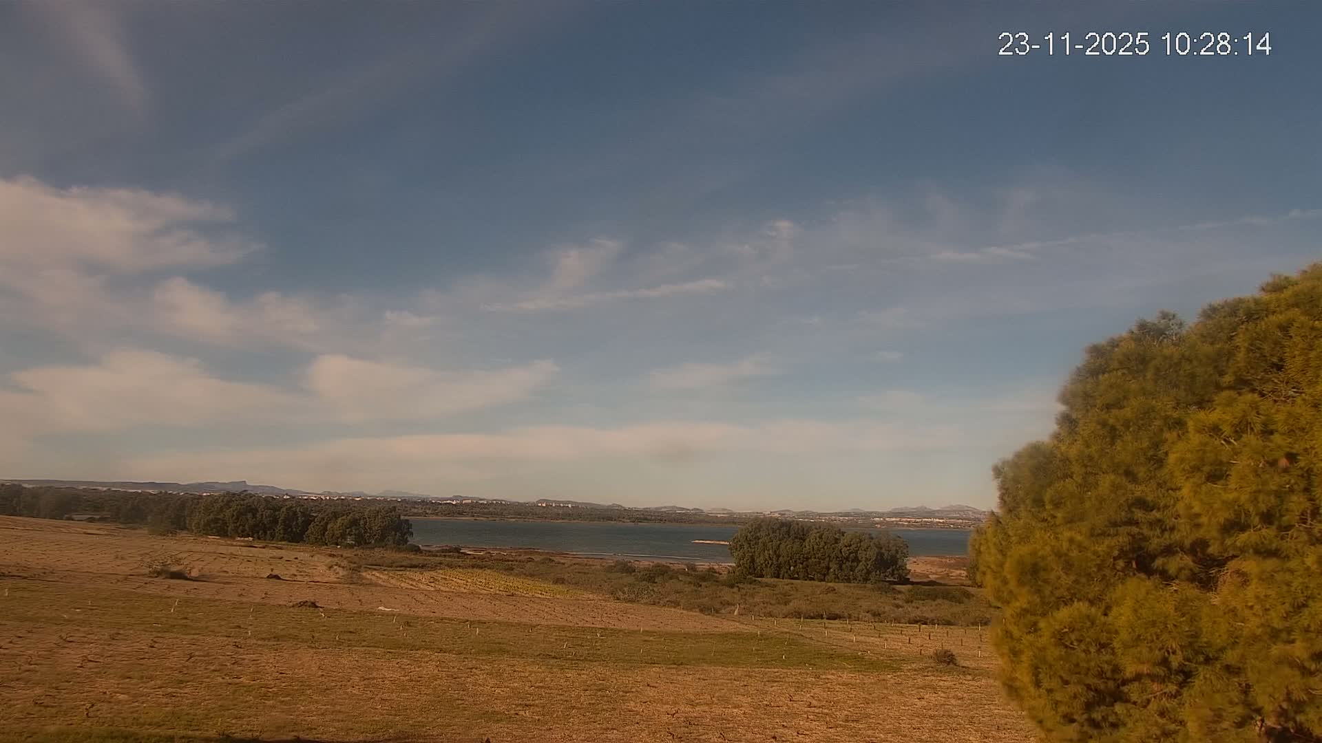 A bright, partly cloudy day illuminates a landscape featuring dry, grassy fields sloping down to a tranquil body of water lined with trees, with distant mountains visible beneath a blue sky.