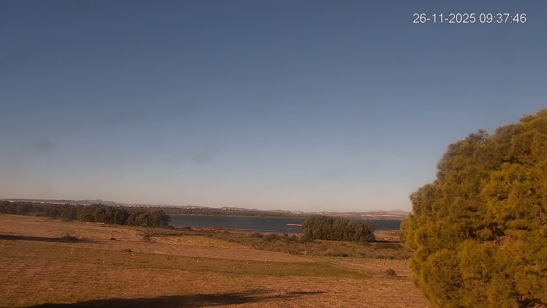 A sun-drenched landscape under a clear blue sky reveals a dry, reddish-brown field in the foreground, leading to a wide body of water flanked by trees and distant hills with a small town on the horizon.