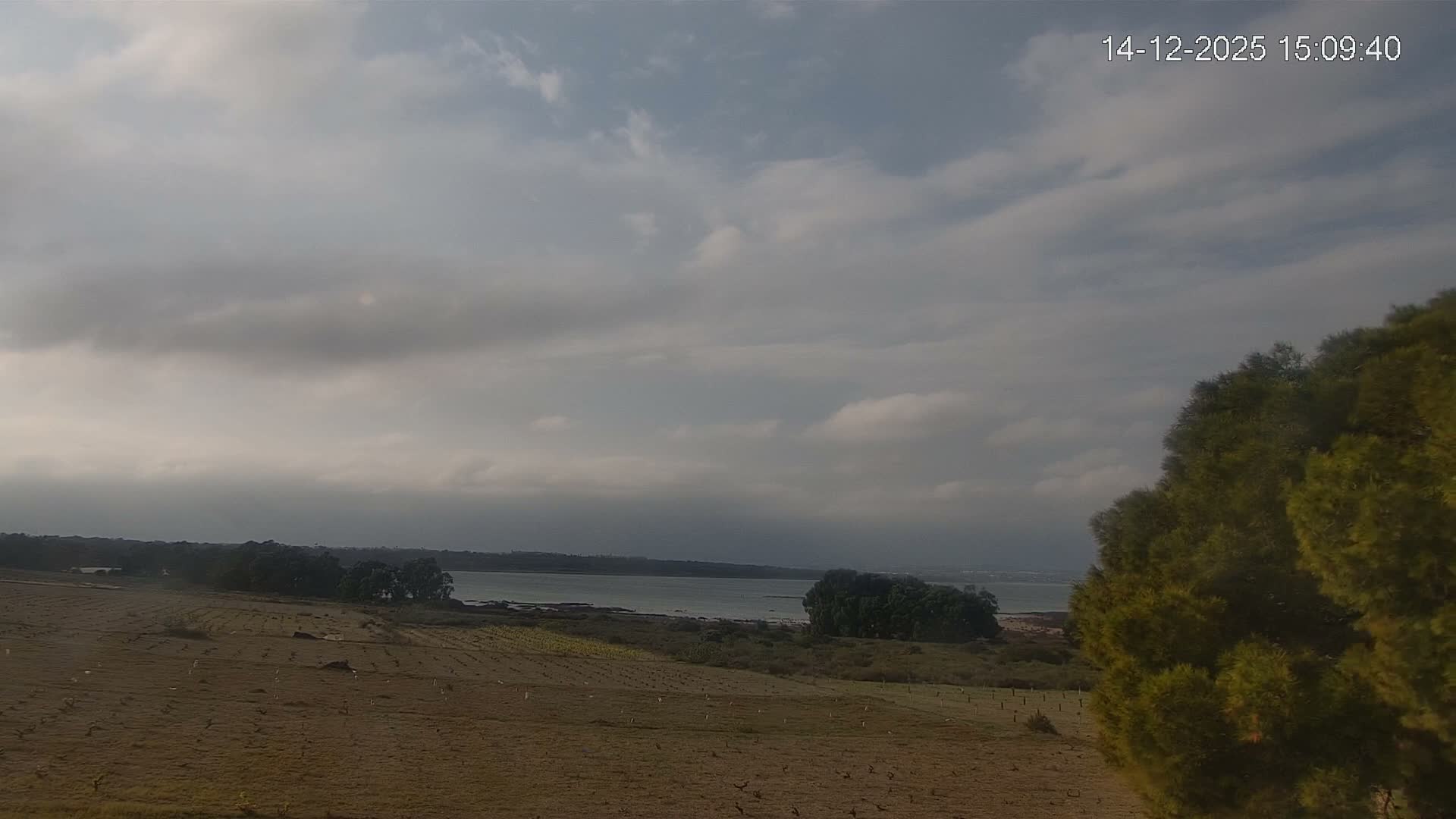 A sun-drenched landscape under a clear blue sky reveals a dry, reddish-brown field in the foreground, leading to a wide body of water flanked by trees and distant hills with a small town on the horizon.