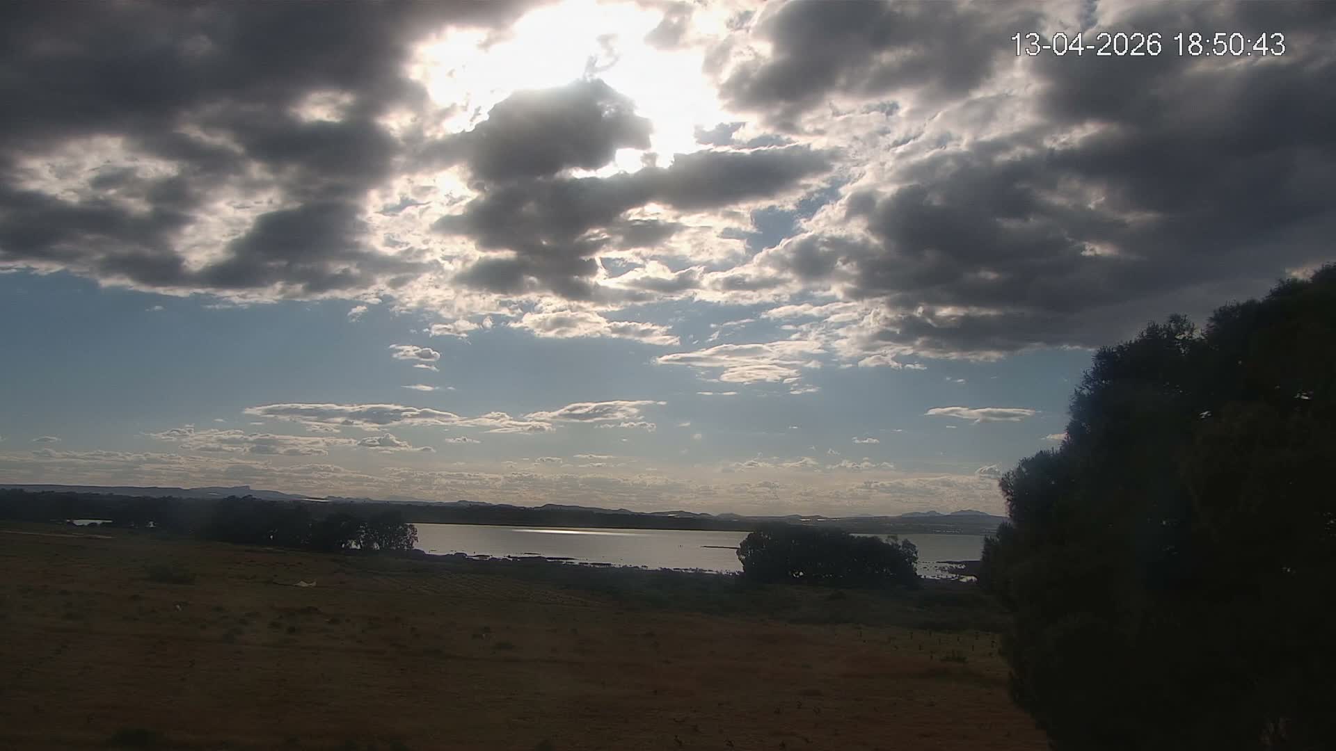 A sun-drenched landscape under a clear blue sky reveals a dry, reddish-brown field in the foreground, leading to a wide body of water flanked by trees and distant hills with a small town on the horizon.