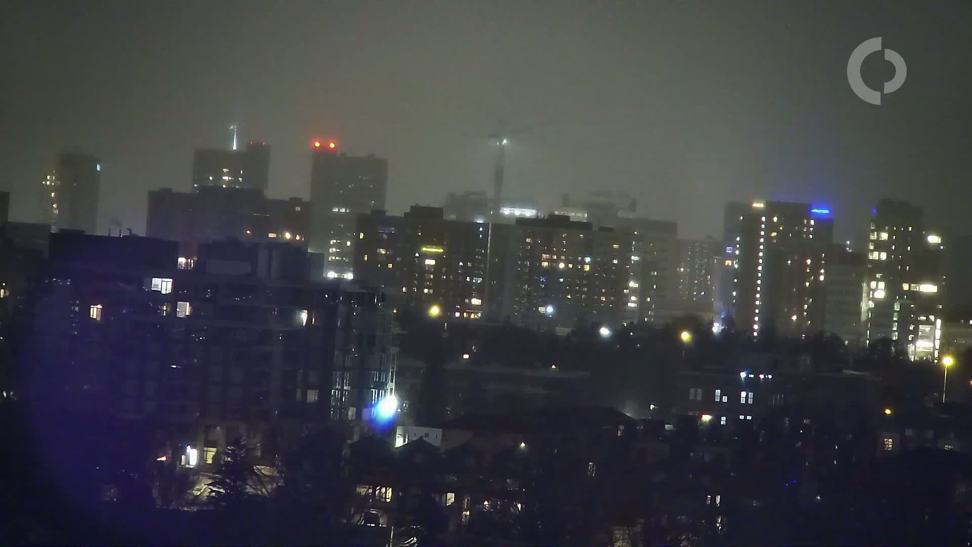 A city skyline at night is shrouded in heavy fog or mist, with numerous apartment buildings illuminated by interior lights.