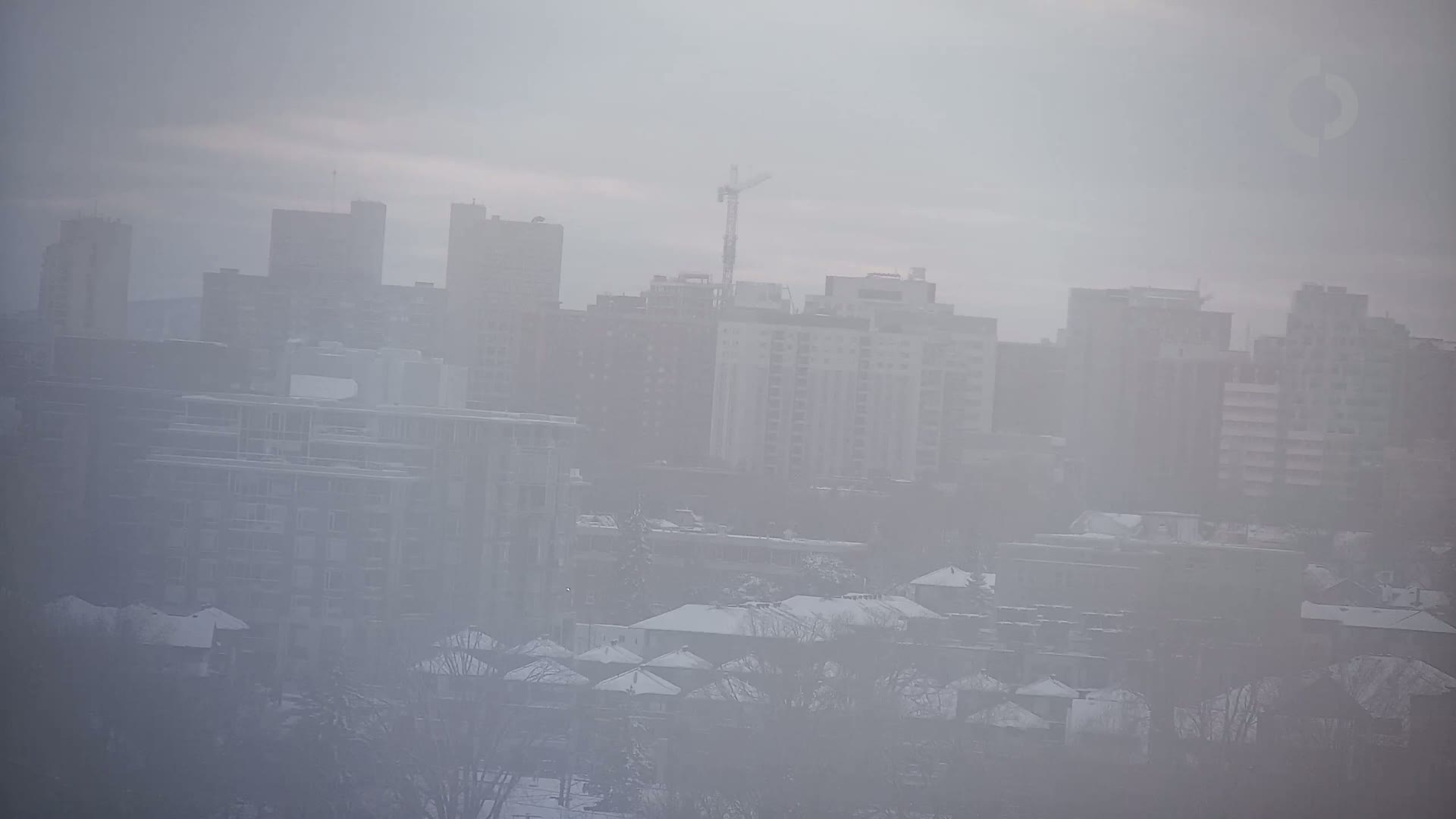 A city skyline is obscured by thick fog, with snow covering the rooftops of houses in the foreground.