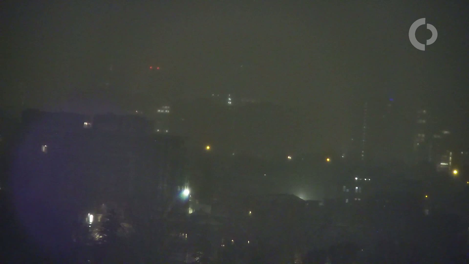 A nighttime cityscape is shrouded in heavy fog, with faint lights visible from buildings and streets.