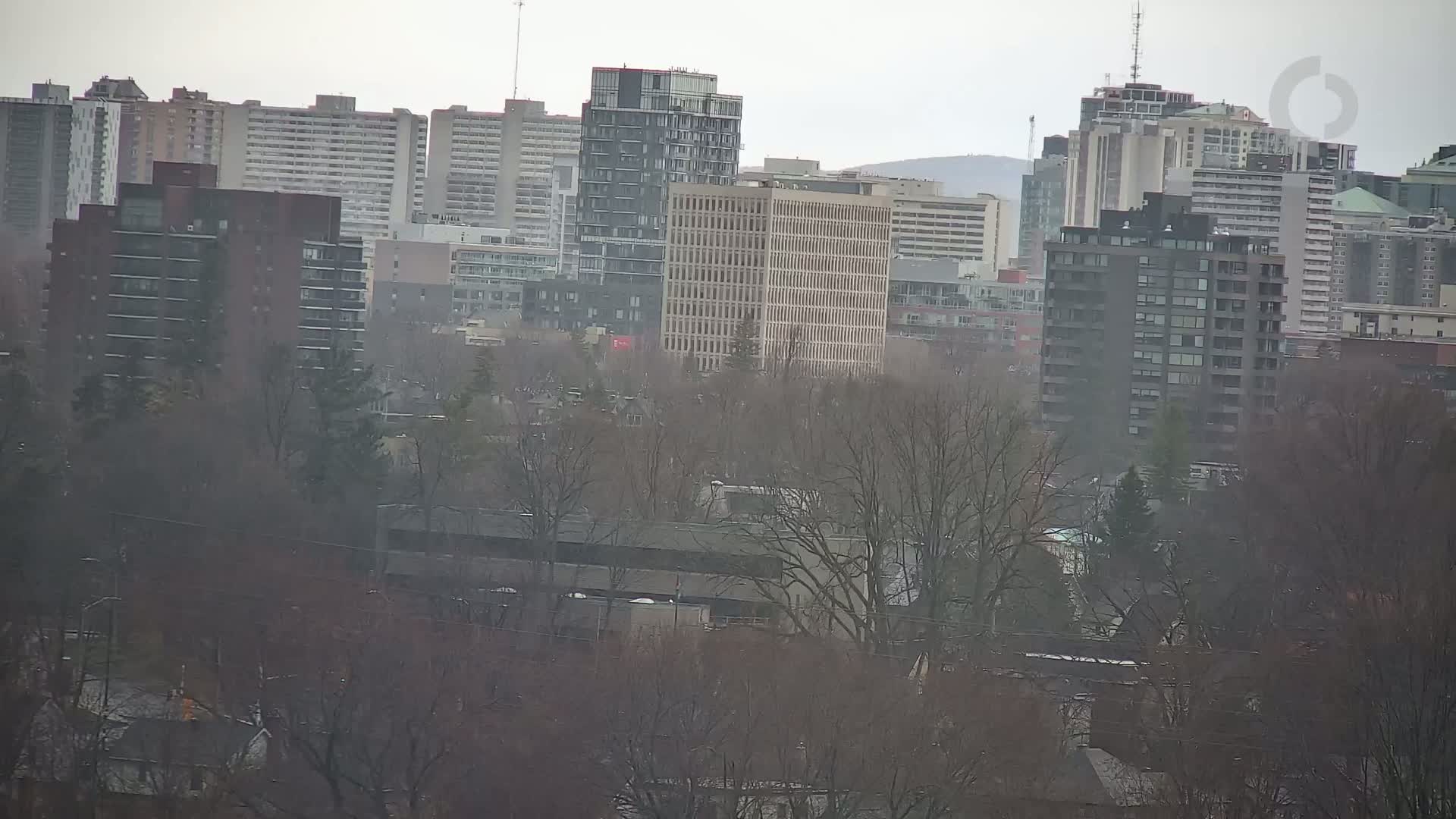 A city skyline with numerous high-rise apartment buildings and trees is visible through an overcast sky.