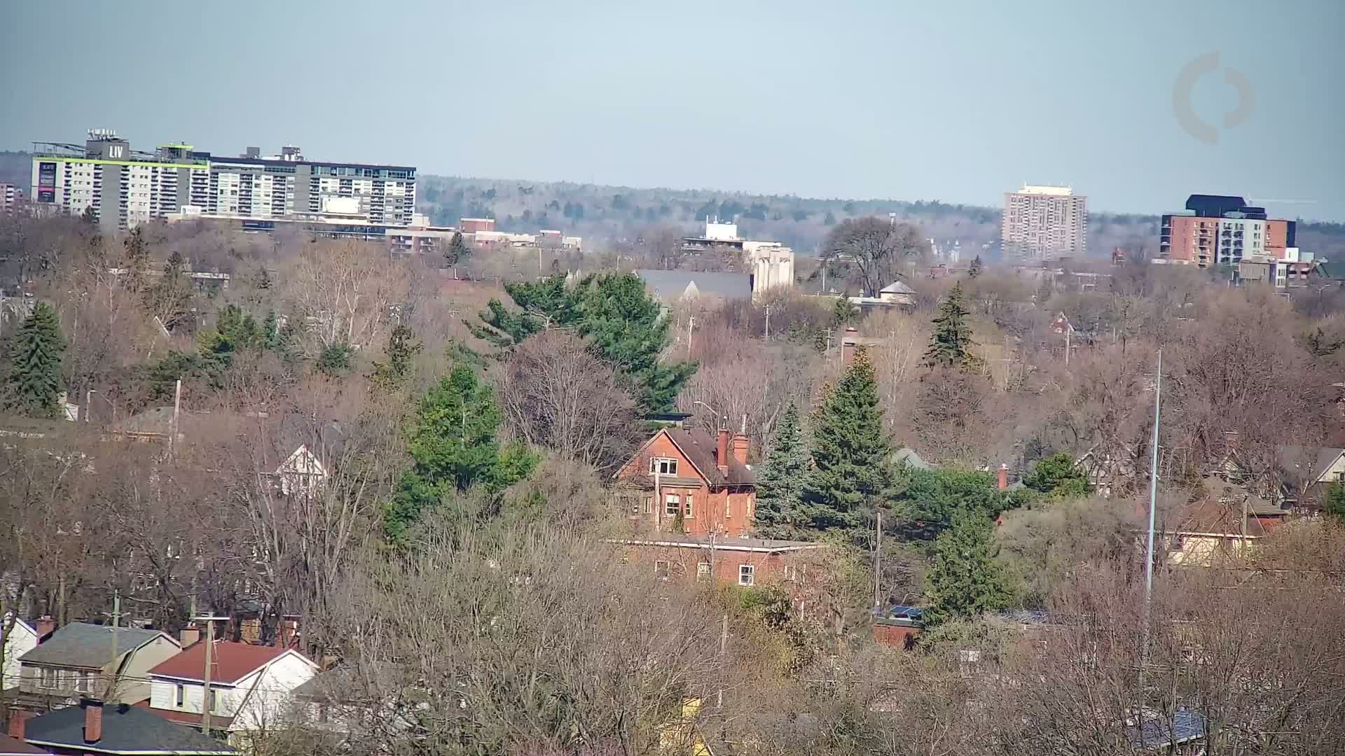 A hazy, sunny day reveals a suburban landscape of houses nestled amongst bare and budding trees, with taller buildings visible in the distance.