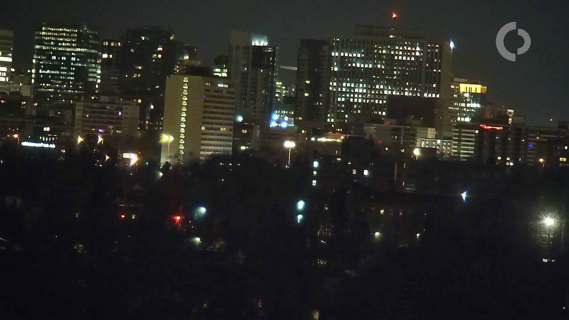 A nighttime cityscape with numerous illuminated buildings, viewed from across a dark expanse of trees and possibly water.