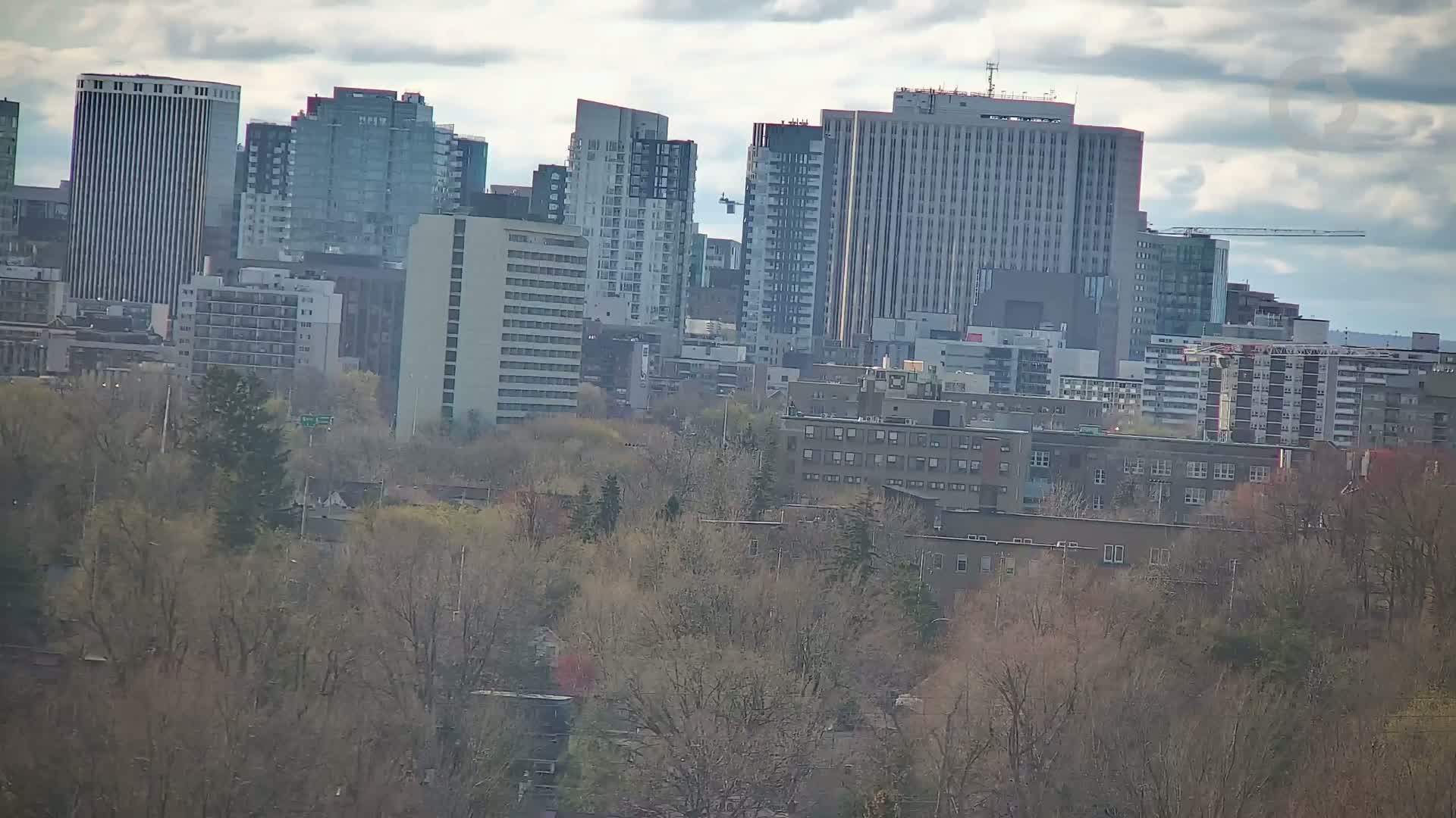 A cityscape of numerous high-rise and low-rise buildings is partially obscured by bare trees under a cloudy sky.