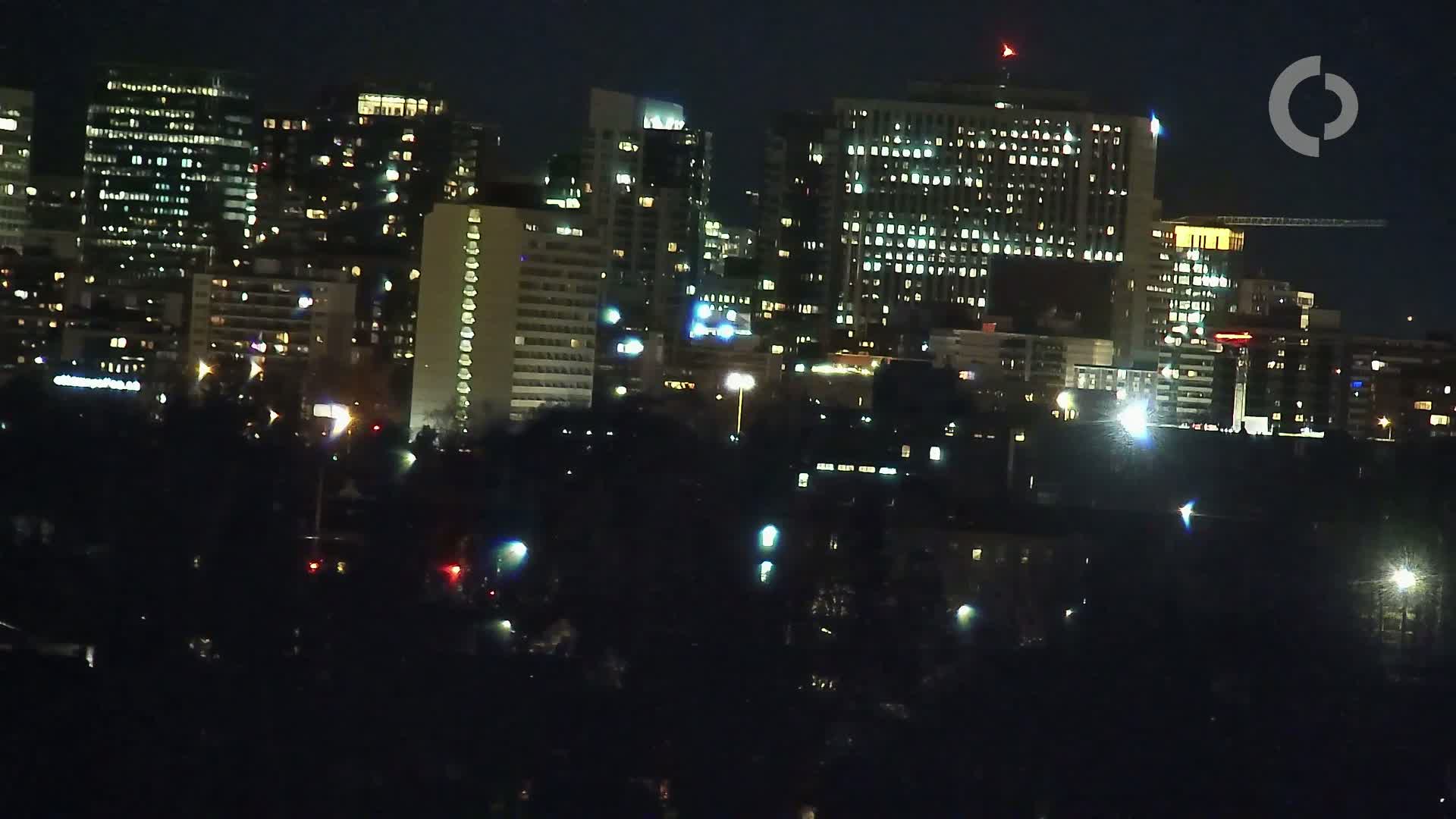 A nighttime cityscape shows numerous illuminated buildings of varying heights, partially obscured by dark trees in the foreground.